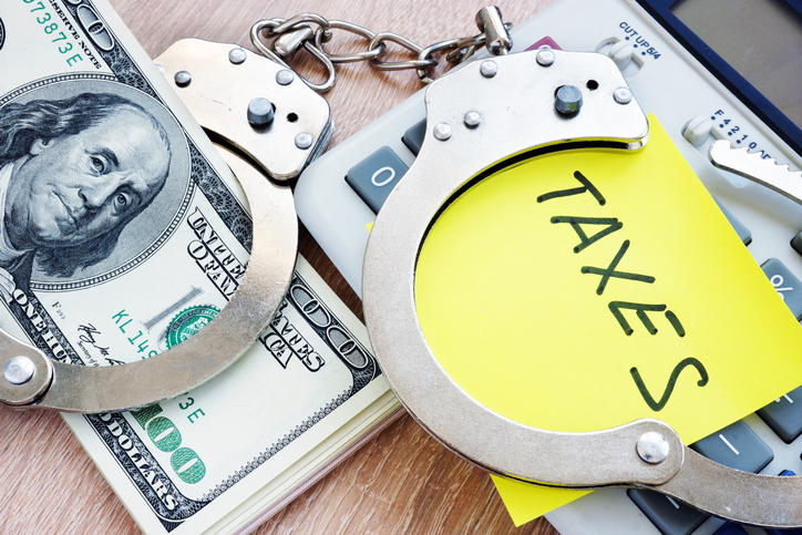 Handcuffs over $100 bills and a yellow post it with the word Taxes written on it.
